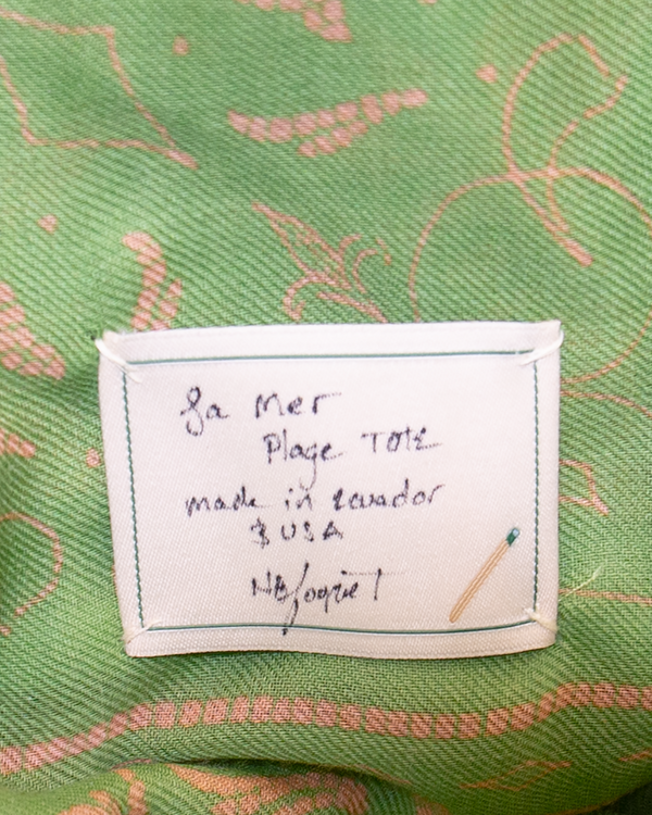 A Mer Vert white fabric label sewn onto green patterned fabric, featuring handwritten text that reads La mer Plage TOTE made in Ecuador USA and a small embroidered matchstick, evoking coastal ease.