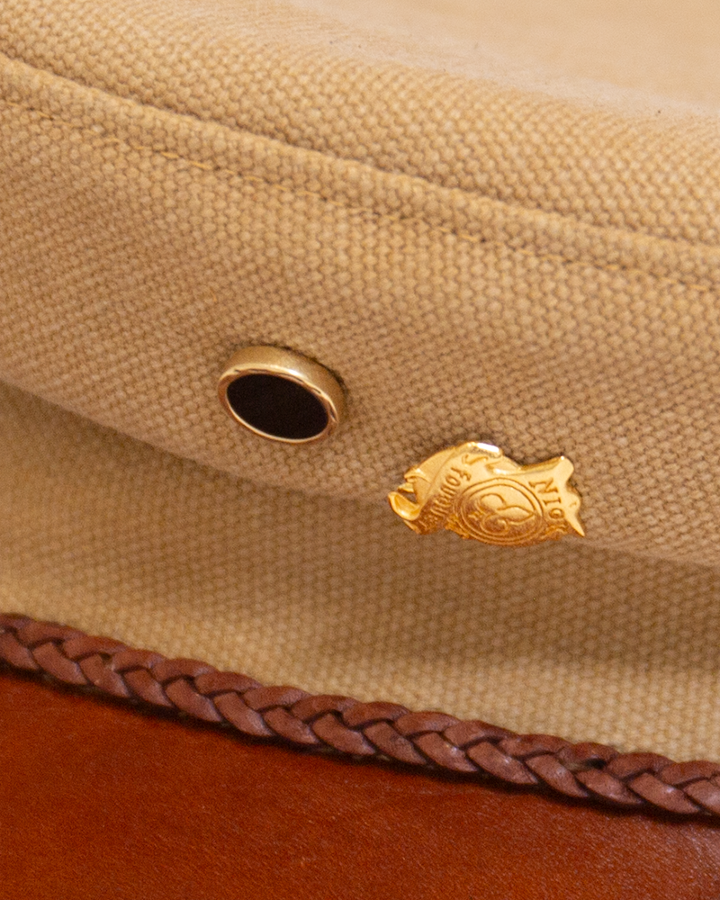 Close-up of the Alcyon tan canvas hat, featuring a black eyelet, a sand-cast jewelry pin, and a braided brown trim along the vachetta brim.