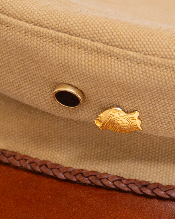 Close-up of the Alcyon tan canvas hat, featuring a black eyelet, a sand-cast jewelry pin, and a braided brown trim along the vachetta brim.